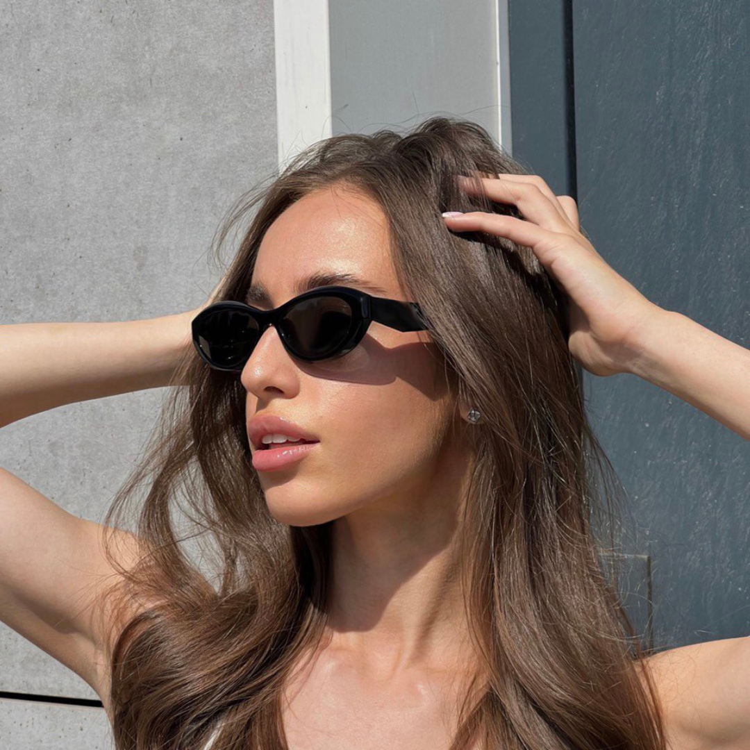 Woman wearing Liora cat eye shades background in Black acetate frame with black TAC lenses, 100% UV protection, modern concrete wall and blue-gray metal door background, looking to the left with both hands intertwined in hair
