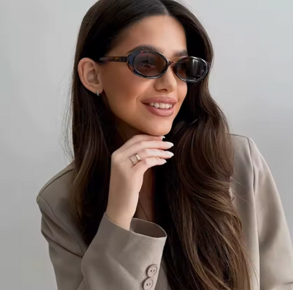 Woman wearing Amélie oval shades in Tortoise polycarbonate frame with amber acrylic lenses, 100% UV protection, white wall background, face and eyes slightly turned to the right with left hand touching chin