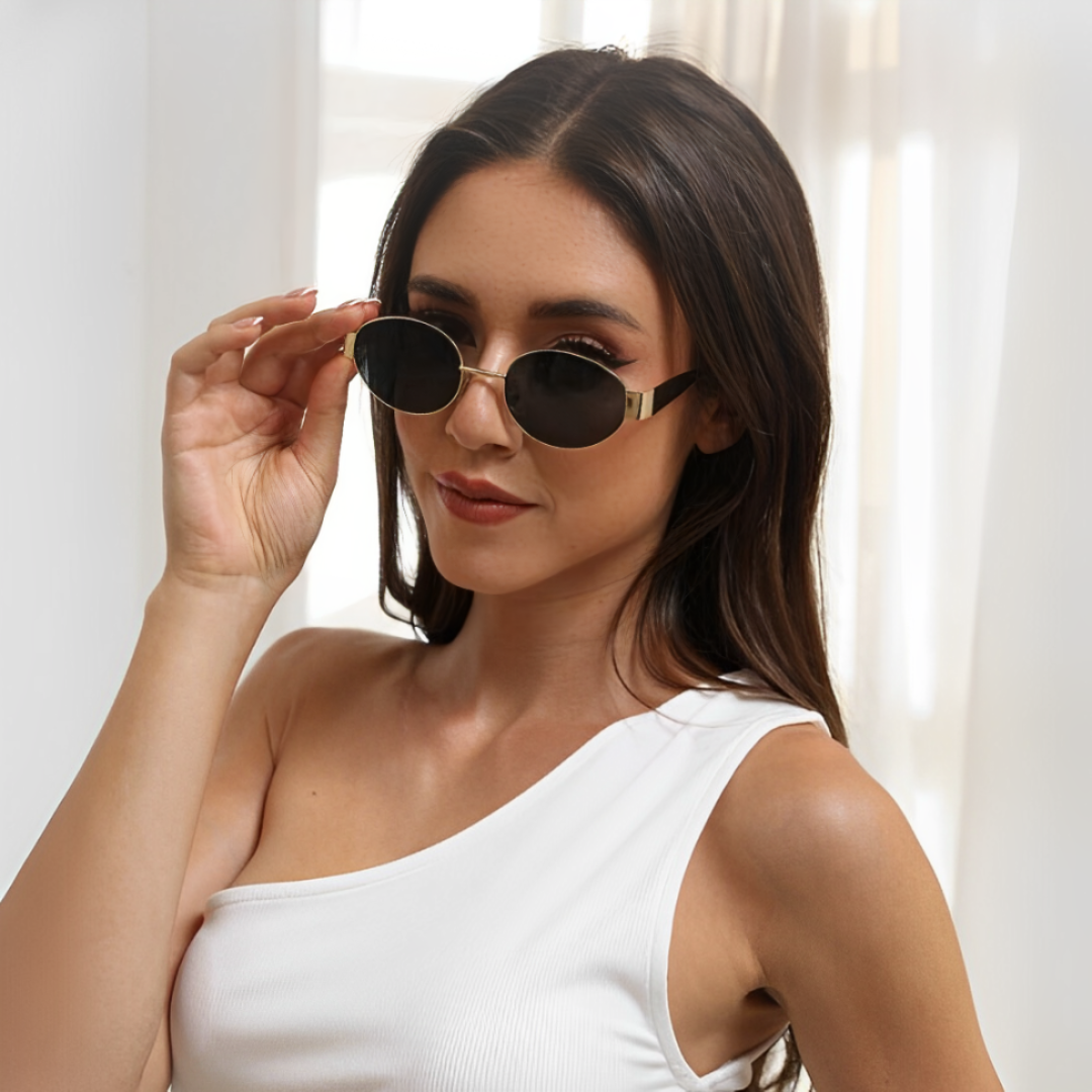 Female model wearing Aurora women’s retro oval shades in Gold frame with Black lenses, against white curtain background, head slightly turned left, looking at the camera, left hand touching the left temple of the frame — stainless steel frame, polycarbonate lenses, UV400 protection