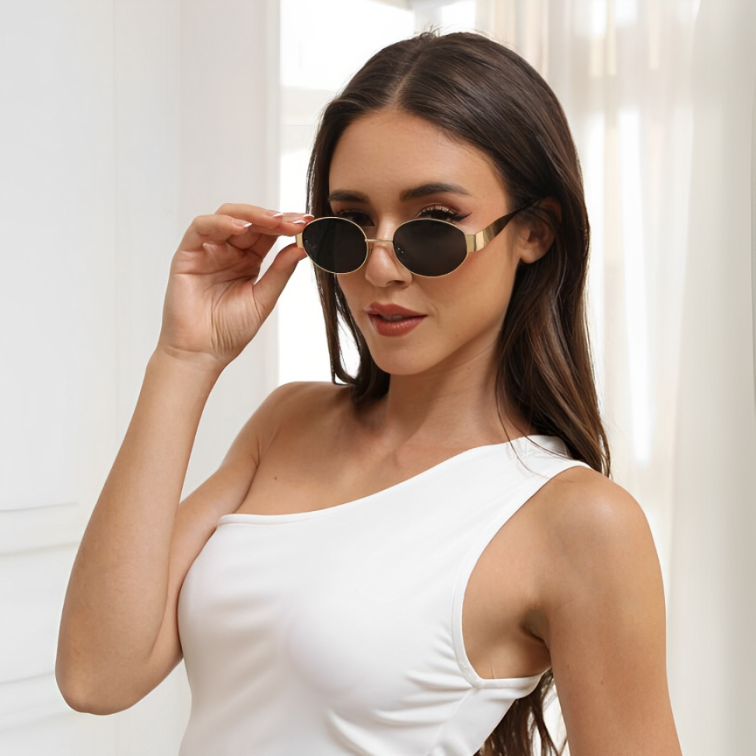 Female model wearing Aurora women’s retro oval sunglasses in Gold frame with Black lenses, against white curtain background, head slightly turned left, looking at the camera with a soft smile, left hand touching the left temple of the frame — stainless steel frame, polycarbonate lenses, UV400 protection