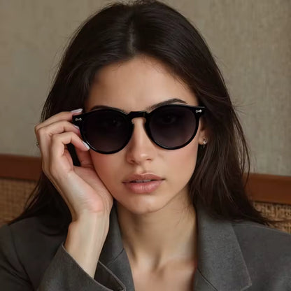 Female model wearing Elysian women’s round sunglasses in Black frame with Black lenses,  against light beige concrete wall with part of a wooden bench visible at the bottom, looking directly at the camera with left hand touching  left side of the sunglasses frame